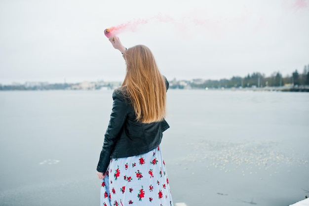 Stijlvol meisje in leren jas houdt roze rookvlam vast op winterdag tegen bevroren meer