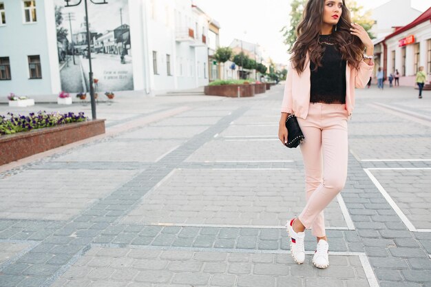 Stijlvol meisje in roze sport chique pak, kanten top en sneakers poseren in de straat met kleine crossbody tas in de hand.