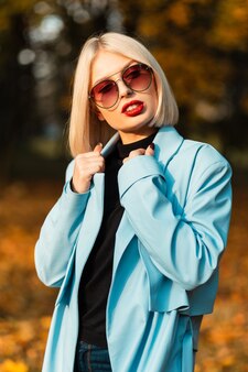 Stijlvol mooi jong meisje met rode lippen en zonnebril in een modieuze blauwe jas met een trui loopt in het park met gouden herfstbladeren