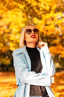 Stijlvol mooi vrouwenmodel met rode lippen in modieuze kleding met vintage zonnebril loopt in een helder herfstpark met geel gebladerte op een zonnige dag