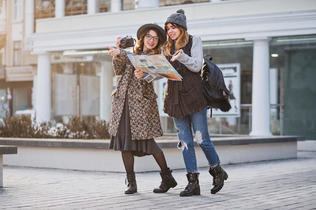 Stijlvol stadsportret van twee modieuze vrouwen die in het moderne stadscentrum van Europa lopen. Modieuze vrienden die reizen met rugzak, kaart, camera, foto maken, toerist, verdwalen, plek voor tekst.