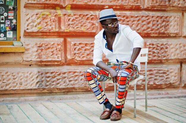 Stijlvolle Afro-Amerikaanse man in wit overhemd en gekleurde broek met hoed en bril poseerde buiten Zwarte modieuze modeljongen