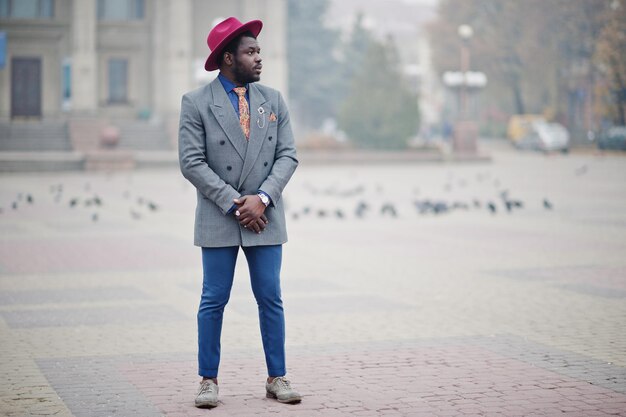 Stijlvolle Afro-Amerikaanse man model in grijze jas stropdas en rode hoed gesteld op straat met mist