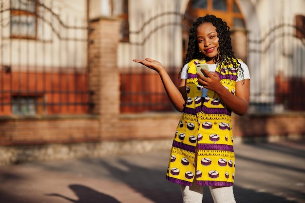 Stijlvolle Afro-Amerikaanse vrouwen in gele jas poseren op straat op zonnige dag met mobiele telefoon bij de hand