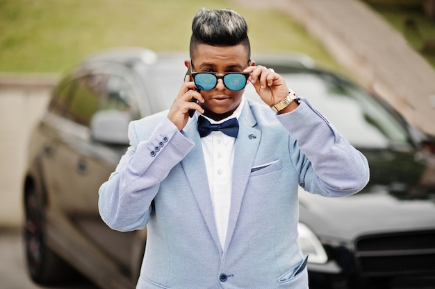 Gratis foto stijlvolle arabische man in jas vlinderdas en zonnebril tegen zwarte suv-auto arabische rijke zakenman die op mobiele telefoon spreekt