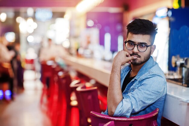 Stijlvolle aziatische man draagt spijkerbroek en bril tegen de bar in de club.