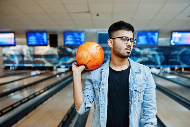 Stijlvolle baard aziatische man in jeans jasje en bril staan op bowlingbaan met bal bij de hand