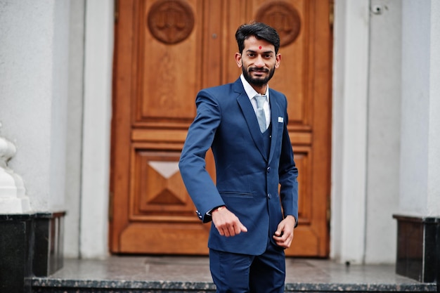 Stijlvolle baard-indiase man met bindi op voorhoofdslijtage op blauw pak buiten tegen de deur van het gebouw