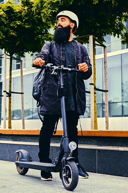 Stijlvolle bebaarde man in zonnebril poseren op elektrische scooter in meer dan modern gebouw achtergrond.