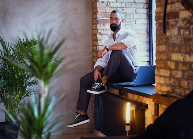 Stijlvolle bebaarde man werkt met een laptop in een kamer met loft-interieur.