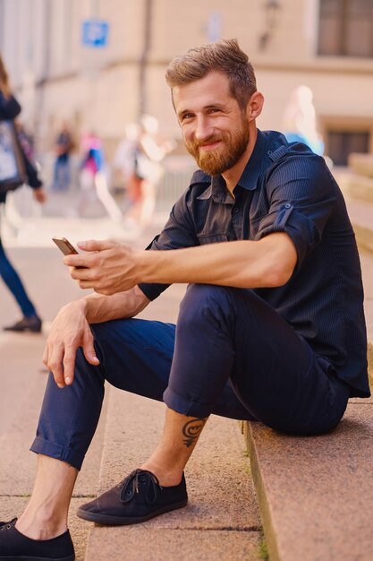 Stijlvolle bebaarde positieve man in een jeans en spijkerjasje zit op een stap en gebruikt een smartphone.