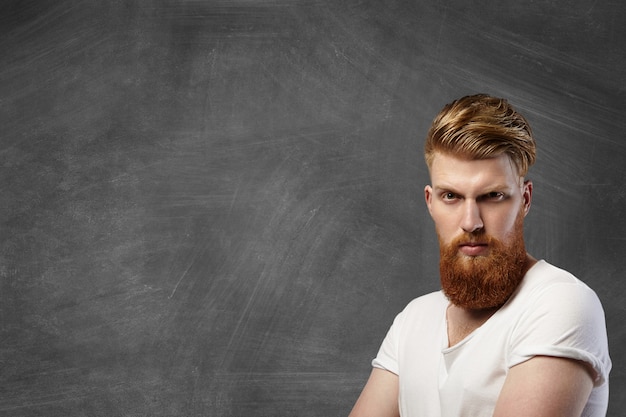 Stijlvolle blanke man met dikke rode baard en hipster kapsel poseren met ernstige brute uitdrukking op zijn gezicht in de rechterbenedenhoek tegen leeg bord met kopie ruimte voor uw inhoud