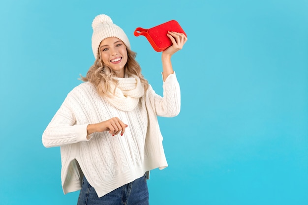 Stijlvolle blonde lachende mooie jonge vrouw met draadloze luidspreker luisteren naar muziek dragen witte trui en gebreide muts poseren op blauw