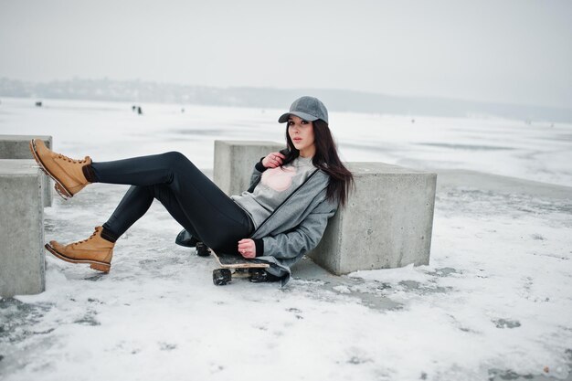 Stijlvolle brunette meisje in grijze pet casual street style met skate board op winterdag