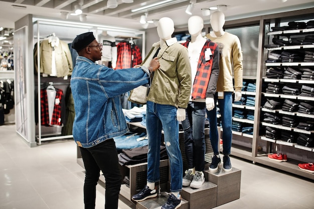 Stijlvolle casual Afro-Amerikaanse man bij jeansjasje en zwarte baret bij kledingwinkel die op nieuwe jas op mannequin kijkt