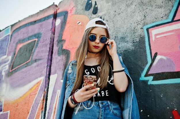 Stijlvolle casual hipster meisje in pet zonnebril en spijkerbroek draagt luistermuziek van hoofdtelefoons van mobiele telefoon tegen grote graffiti muur