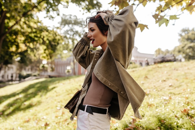 Stijlvolle dame met brunette kort kapsel in spijkerjasje vormt buitenshuis. koele vrouw die in witte jeans weg naar buiten kijkt.