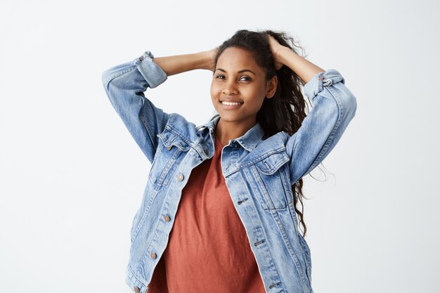 Stijlvolle donkere tiener met golvend haar met vrolijke glimlach. Mooi jong wijfje in denimjasje en rode t-shirt die haar haar vegen dat op wit wordt geïsoleerd.