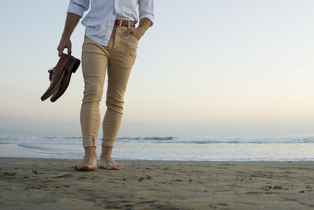 Stijlvolle elegante man in wit overhemd en beige broek wandelen op het strand met zijn schoenen in handen