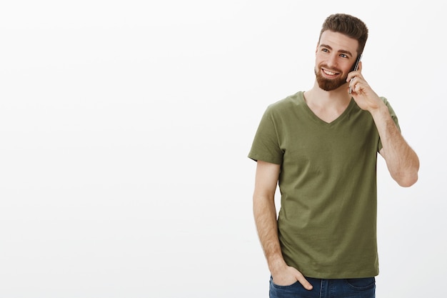 stijlvolle en zelfverzekerde knappe bebaarde mannelijke ondernemer die op smartphone lacht en lacht genietend van gesprek kijkend naar linkerbovenhoek over witte muur