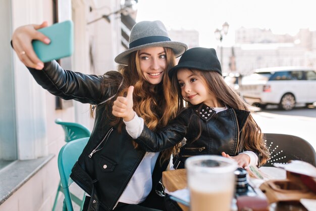 Stijlvolle familie zittend in een stadscafé, kijk in de telefoon en neem selfies en glimlach op de zonnige stadsachtergrond. Het meisje toont vinger aan de bovenkant die camera bekijkt. Echte emoties, goed humeur.