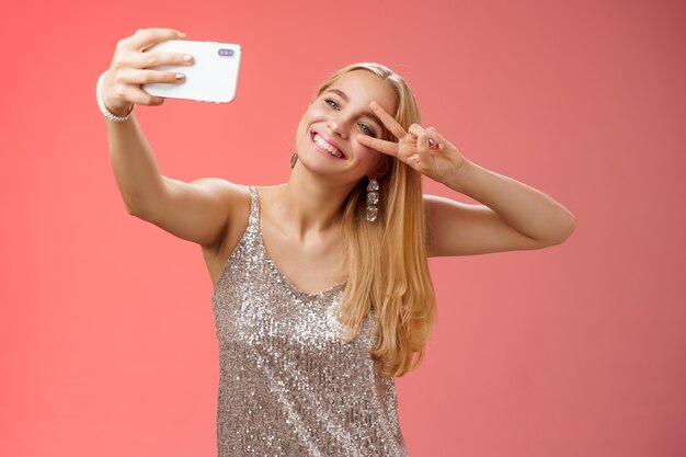 Stijlvolle fantastische glamour jonge blonde vrouw in glinsterende zilveren jurk kantelbaar hoofd zorgeloos show vredesgebaar overwinningsteken uitbreiden arm houden smartphone nemen selfie video post online opnemen