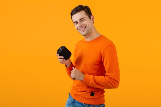 Stijlvolle glimlachende jonge man in oranje trui met draadloze luidspreker die gelukkig naar muziek luistert