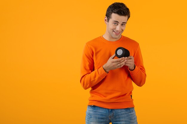 Stijlvolle glimlachende jongeman in oranje trui met draadloze luidspreker die graag naar muziek luistert met plezier