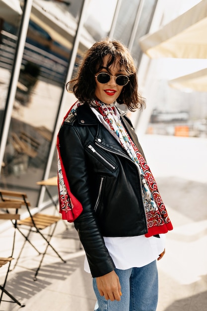 Gratis foto stijlvolle hipster meisje draagt lederen jas met korte haarstijl en rode lippenstift wandelen in de stad in het zonlicht