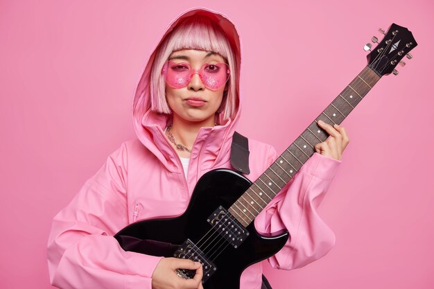 Stijlvolle hipster vrouw draagt trendy zonnebril jas met capuchon poses met zwarte elektrische gitaar ziet er serieus uit, maakt nieuw nummer voor haar album