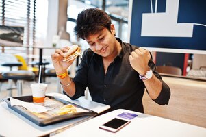 Stijlvolle indiase man zit in fastfoodcafé en eet hamburger lees ochtendnieuws op mobiele telefoon hij is blij en laat zien dat hij iets wint