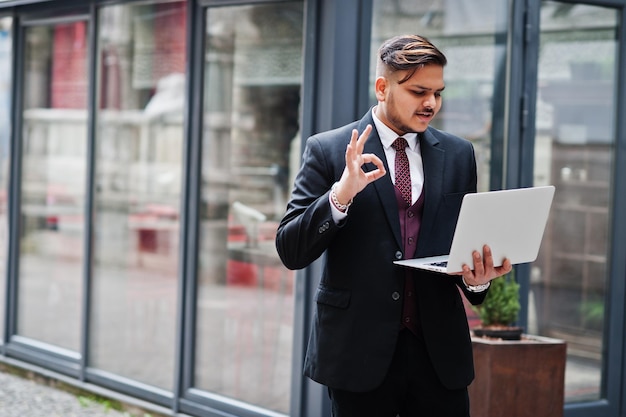 Stijlvolle indiase zakenman in formele kleding met laptop op handen tegen ramen in zakencentrum toon ok teken met vingers