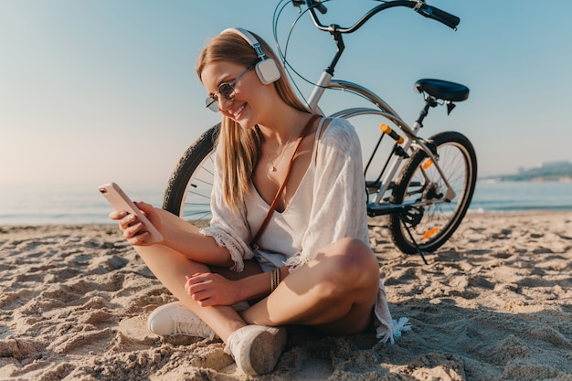 Stijlvolle jonge aantrekkelijke blonde lachende vrouw zittend op het strand met fiets in koptelefoon luisteren naar muziek