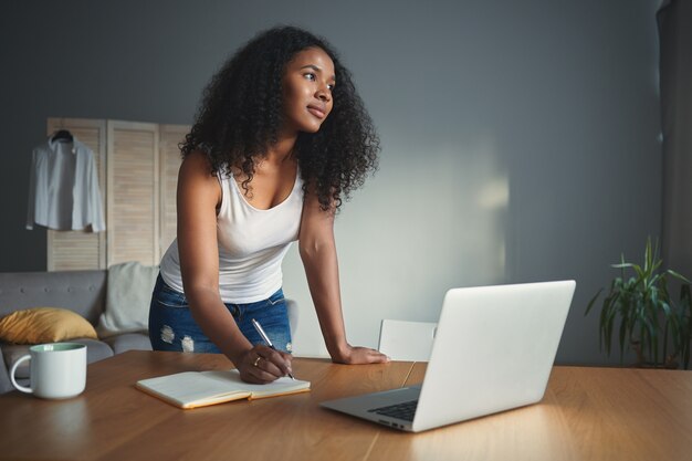 Stijlvolle jonge Afro-Amerikaanse vrouw journalist met krullend haar permanent aan bureau met geopende laptop en opschrijven in voorbeeldenboek, onderzoek doen voor nieuw artikel. Mensen, beroep en technologie