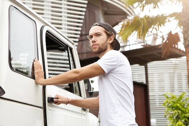 Stijlvolle jonge bebaarde man in snapback die hand op handvat van zijn witte jeep houdt, deur opent terwijl hij instapt, weekend gaat doorbrengen op safarirace