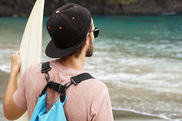 Stijlvolle jonge bebaarde surfer dragen van een zonnebril en snapback kijken naar blauwe zee golven terwijl je op zandstrand met zijn bodyboard. sport, extreem, hobby