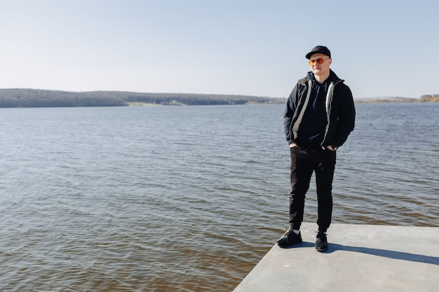 Stijlvolle jonge jongen in oranje bril, jas en pet, loop rond het meer in de lente of herfst