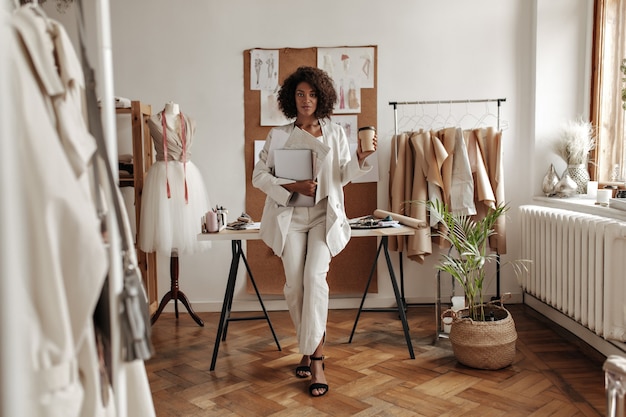 Stijlvolle jonge krullende donkere vrouw in witte broek, jas en blouse leunt op bureau in modeontwerper kantoor, houdt koffiekopje en laptop