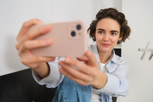 Gratis foto stijlvolle jonge meidenblogger neemt selfie in haar keuken poseert voor foto op mobiele telefoon met behulp van camera