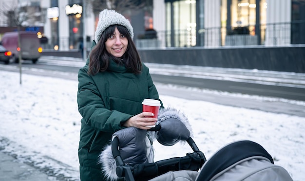 Stijlvolle jonge moeder met een kopje koffie op een wandeling met een kinderwagen in de winter