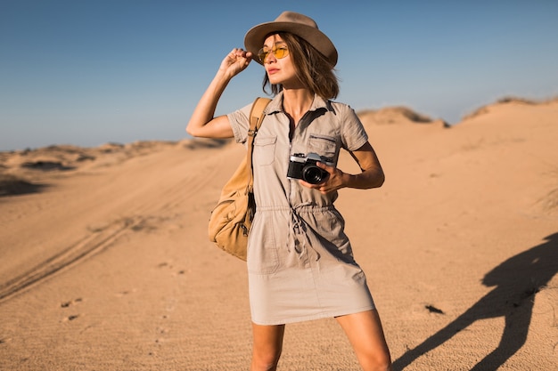 Stijlvolle jonge vrouw in kaki jurk wandelen in de woestijn, reizen in Afrika op safari, hoed en rugzak dragen, foto nemen op vintage camera