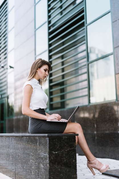 Stijlvolle jonge zakenvrouw zittend op de bank met behulp van laptop in de open lucht