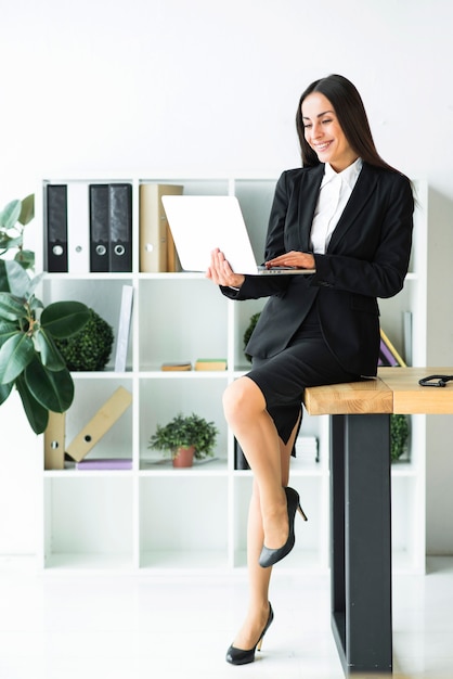 Stijlvolle jonge zakenvrouw zittend op een bureau met behulp van laptop