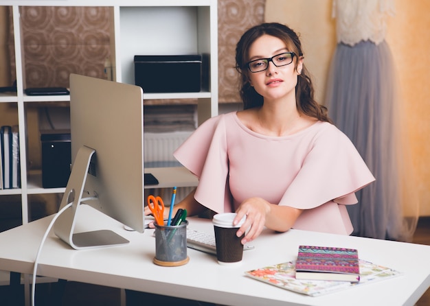 Stijlvolle jongedame in roze luxe jurk werken op kantoor op computer