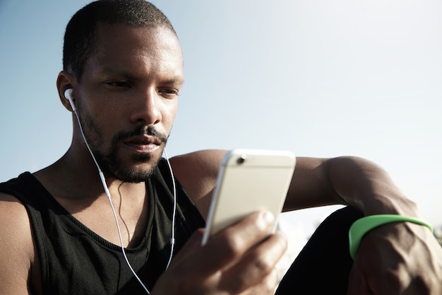 Gratis foto stijlvolle jongere zittend op de trap bij het water en luisteren naar muziek. eenzame afro-amerikaanse man in zwarte mouwloos met groene fitness tracker genieten van muziek en sms'en op het apparaat.