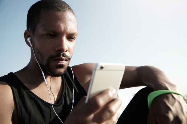 Stijlvolle jongere zittend op de trap bij het water en luisteren naar muziek. Eenzame Afro-Amerikaanse man in zwarte mouwloos met groene fitness tracker genieten van muziek en sms'en op het apparaat.