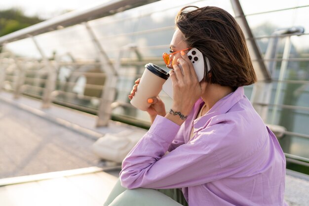 Stijlvolle kortharige vrouw zittend op de moderne brug, genieten van koffie en het gebruik van mobiele telefoon