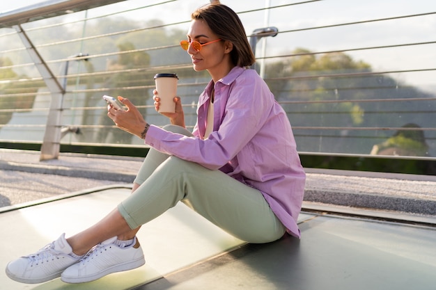 Stijlvolle kortharige vrouw zittend op de moderne brug, genieten van koffie en het gebruik van mobiele telefoon