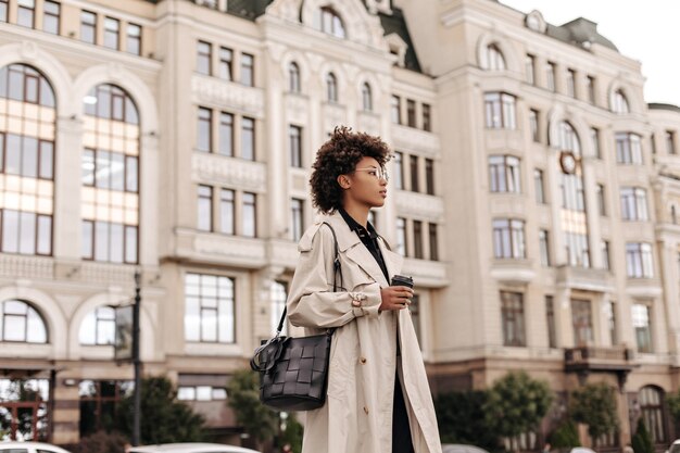 Stijlvolle krullende dame in beige trenchcoat, bril en met zwarte tas houdt koffiekopje vast en loopt in de stad