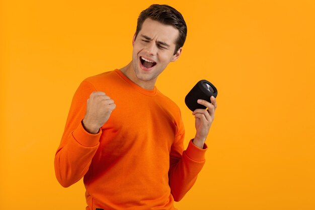 Stijlvolle lachende jonge man in oranje trui met draadloze luidspreker gelukkig luisteren naar muziek plezier op oranje